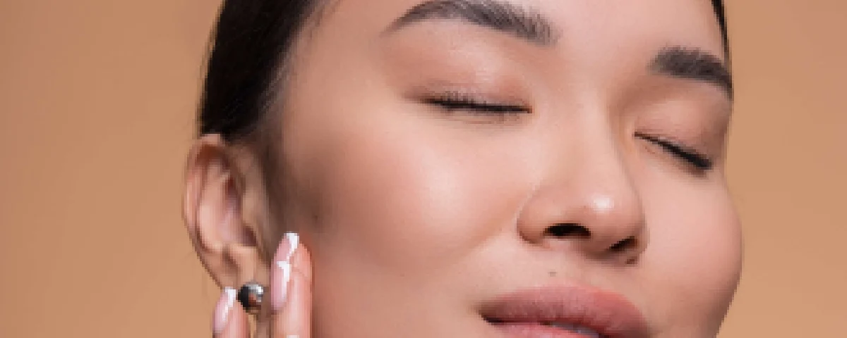 Close-up portrait of an Asian woman with her eyes closed, gently touching her clear, glowing skin after Facial Treatments in Salem, NH, against a beige background.