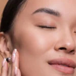 Close-up portrait of an Asian woman with her eyes closed, gently touching her clear, glowing skin after Facial Treatments in Salem, NH, against a beige background.