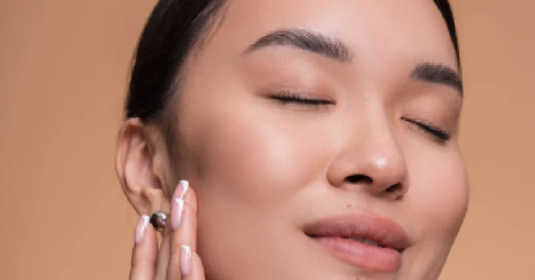 Close-up portrait of an Asian woman with her eyes closed, gently touching her clear, glowing skin after Facial Treatments in Salem, NH, against a beige background.