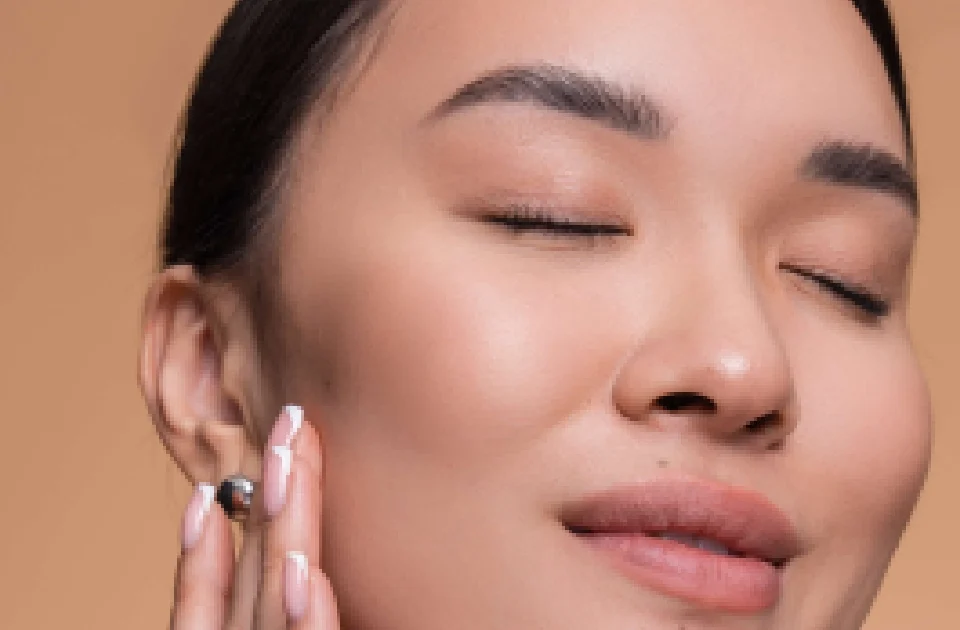 Close-up portrait of an Asian woman with her eyes closed, gently touching her clear, glowing skin after Facial Treatments in Salem, NH, against a beige background.